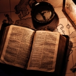 An open bible and compass placed on a map, lit by a lantern.